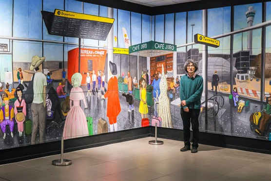 A man wearing a blue jumper and black trousers stands in front on a large mural of an airport
