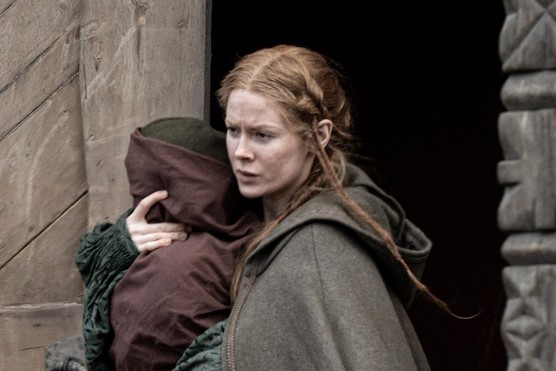 A woman in a grey cloak holds a swaddled infant to her shoulder
