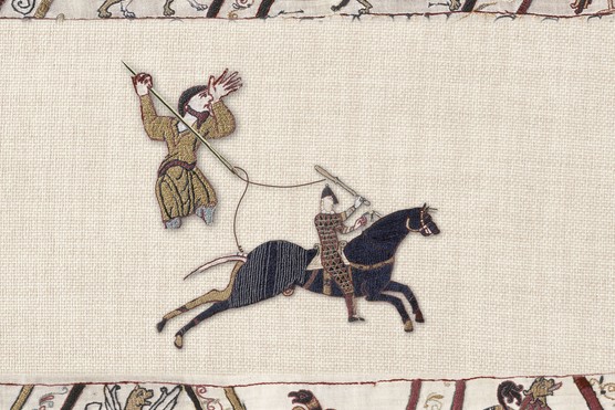 A tapestry scene showing a small figure dressed in a gold robe holding a large needle and thread. Below him, another man rides on the back of a black horse